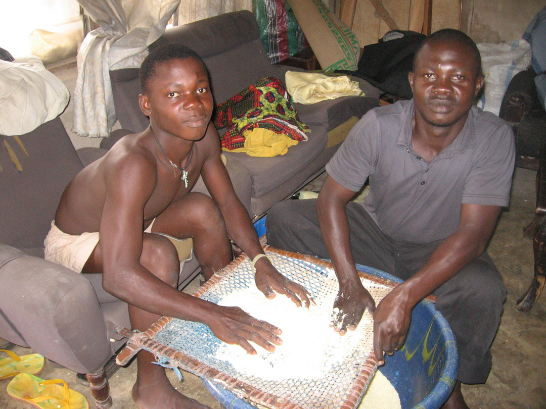 Two young men sift the product of mashed and dried yams through a silt to prepare it for cooking.